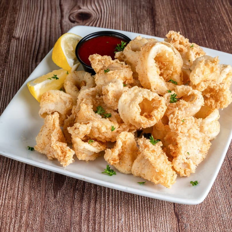 Crispy fried calamari with lemon wedges and a side of red marinara sauce on a white plate.